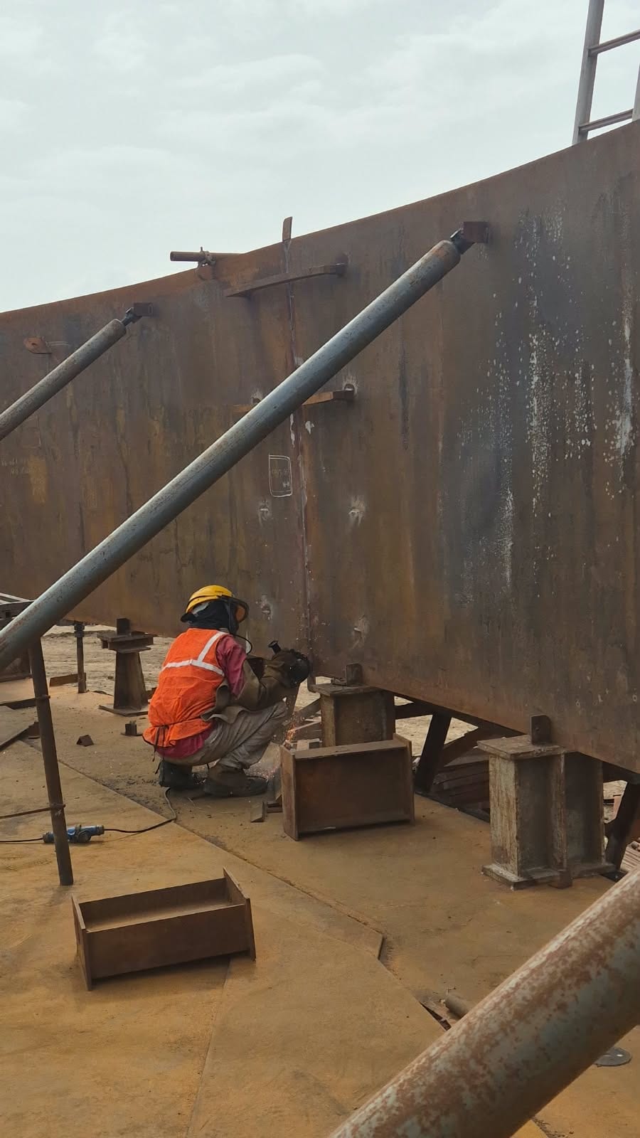 Worker welding a large steel storage-tank shell supported on steel stools